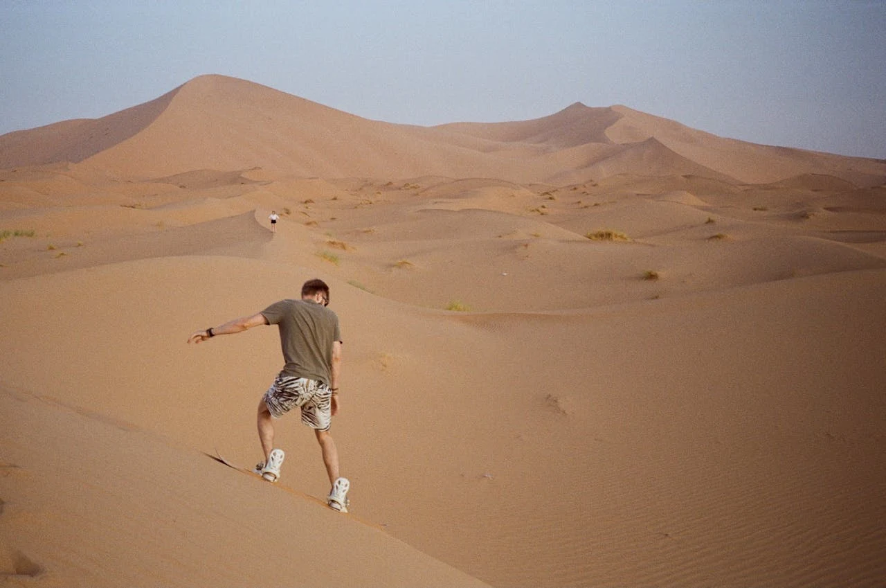 Tourist sandboarding down a large golden dune in Merzouga, action shot, bright sun, sand spray, joyful expression
